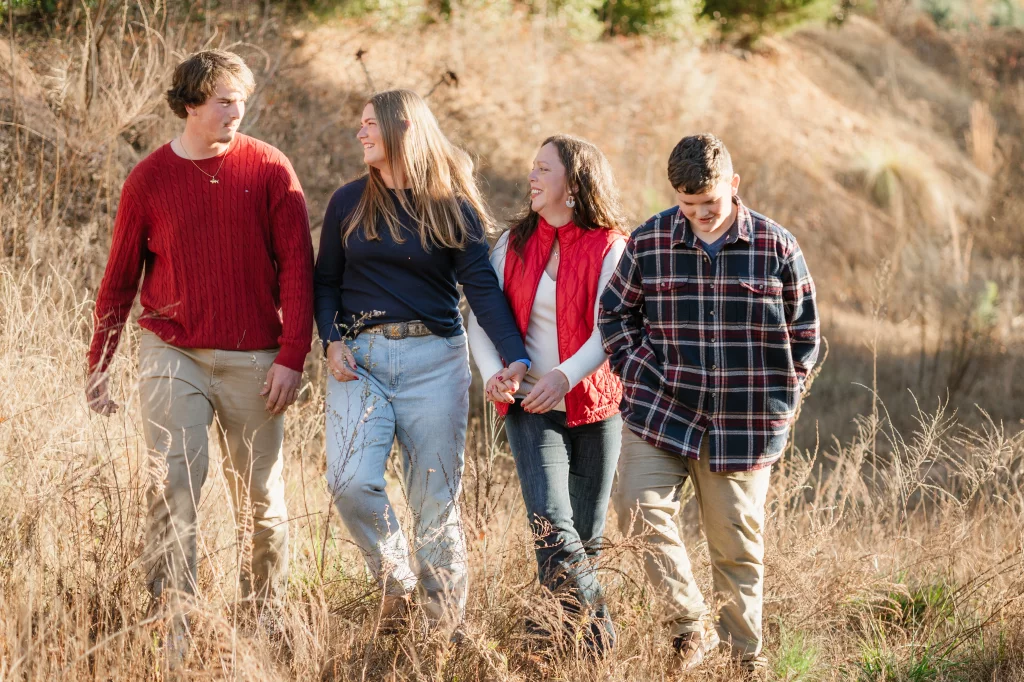 The Garman's family photos taken at BridgeWay Station, Simpsonville: Four people walking together through a dry grassy field, two women in the middle holding hands and smiling at each other, a man in a red sweater on the left and a boy in a plaid shirt on the right.