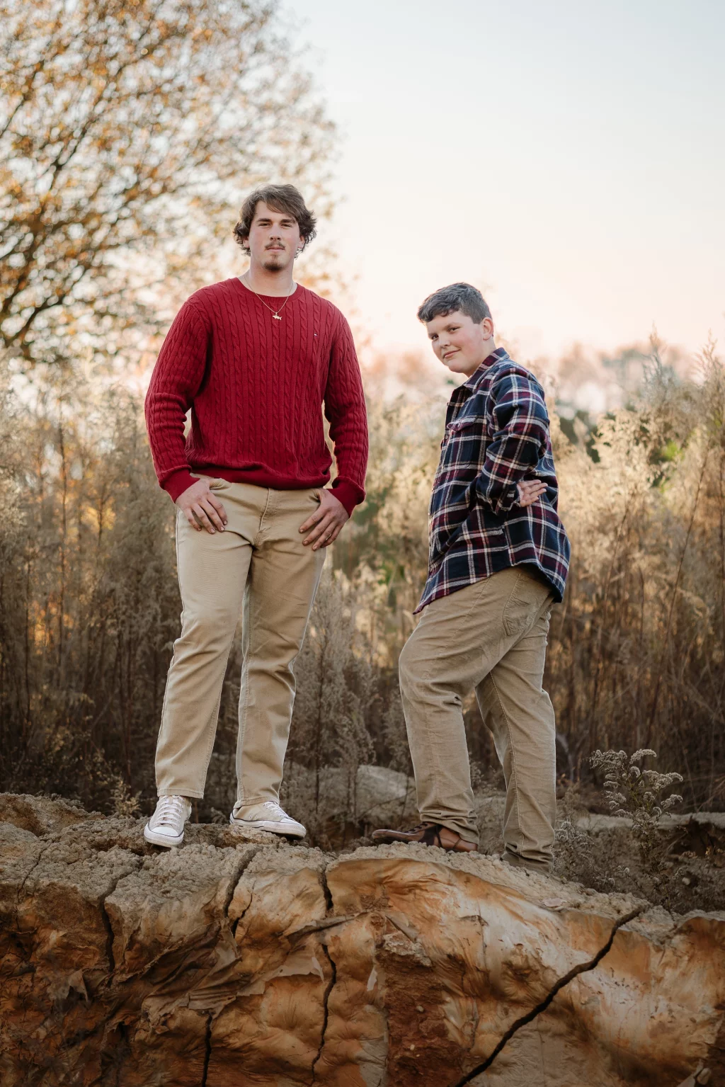 The Garman's family photos taken at BridgeWay Station, Simpsonville: Two males standing on a large rock outdoors, one wearing a red sweater and beige pants, the other wearing a plaid shirt and beige pants, with a background of dry vegetation and a tree.