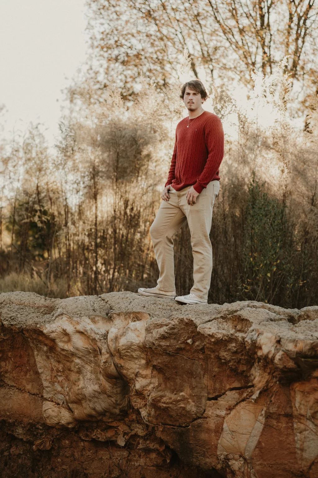 The Garman's family photos taken at BridgeWay Station, Simpsonville: Young man in red sweater and beige pants standing on a rocky ledge with dry trees in the background.