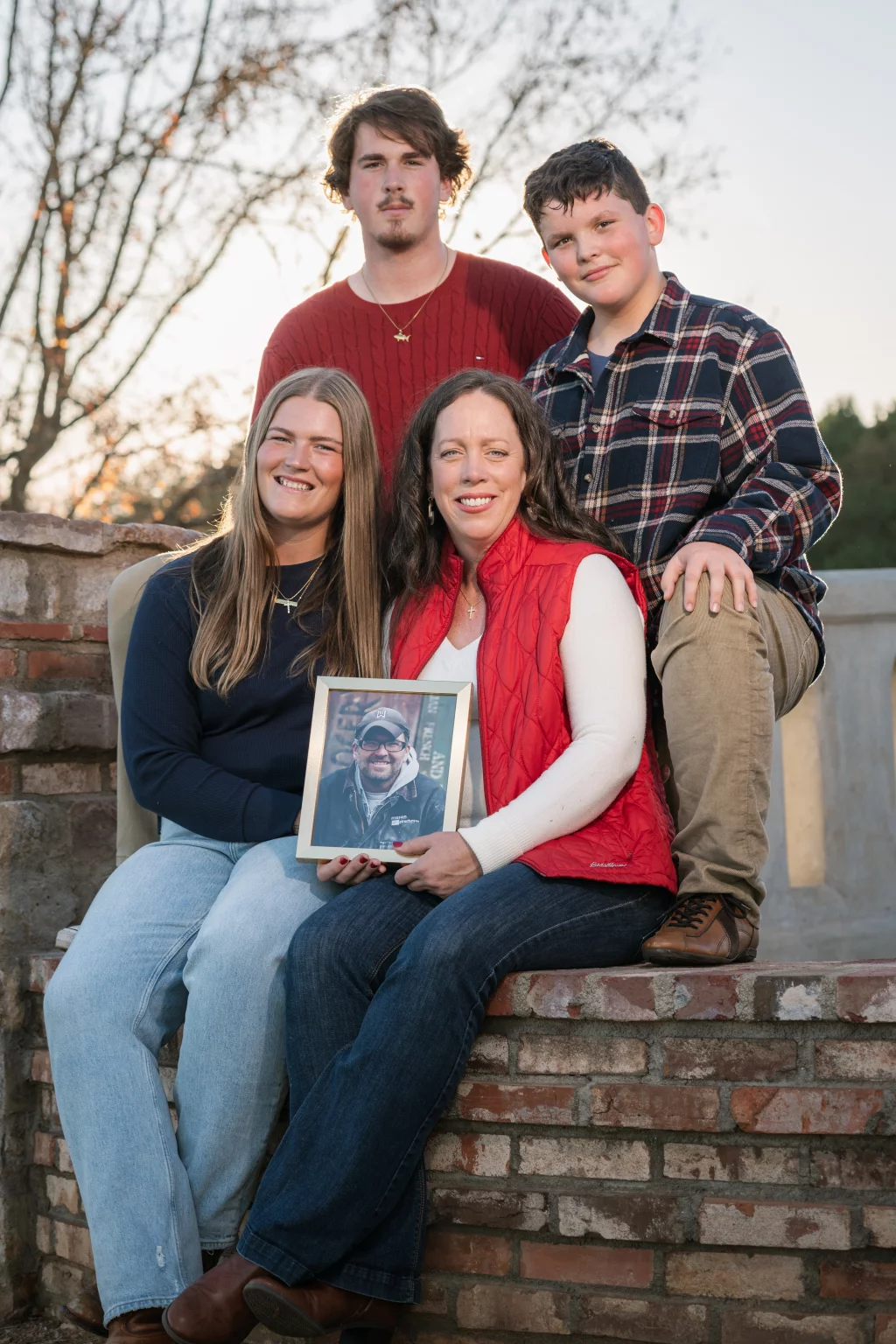 The Garman's family photos taken at BridgeWay Station, Simpsonville: Group of four people sitting and standing on a brick wall outdoors, two women seated in front, one woman holding a framed photo of a smiling man wearing glasses and a cap, with two boys standing behind them.