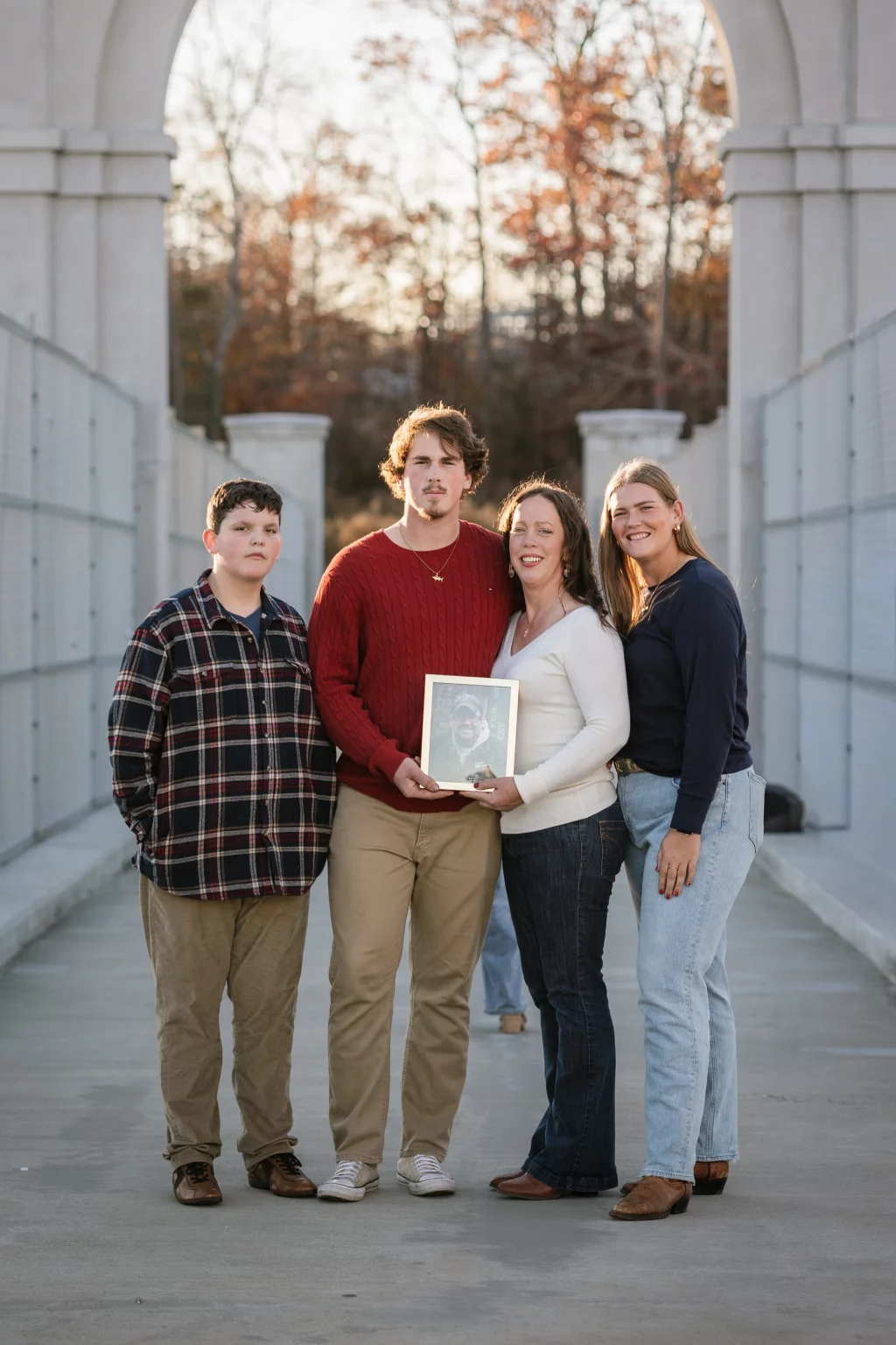 The Garman's family photos taken at BridgeWay Station, Simpsonville: Four people standing together on a bridge under an archway, with three adults and one child; one adult holds a framed photo of a man.