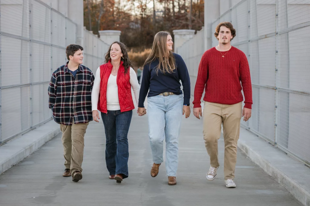 The Garman's family photos taken at BridgeWay Station, Simpsonville: Four people walking side by side on a paved path between metal fences, two women in the center holding hands, with autumn trees in the background.