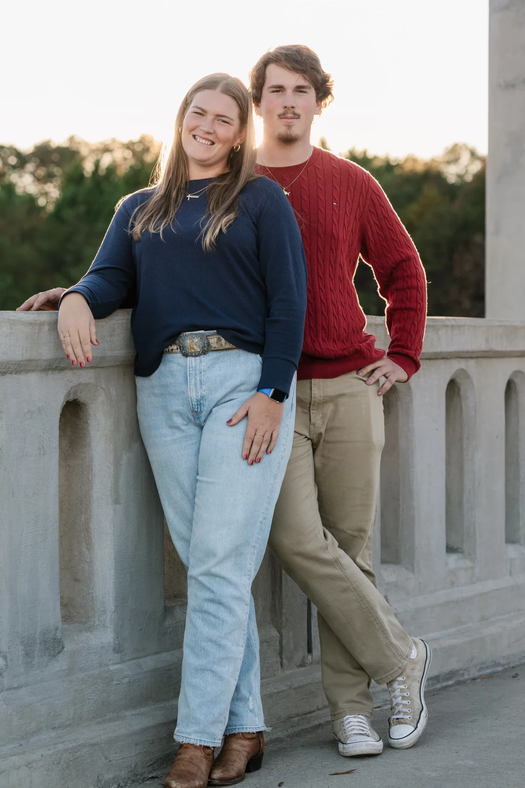 The Garman's family photos taken at BridgeWay Station, Simpsonville: Young woman and man standing outdoors by a stone railing, smiling, with trees and sunset in the background.