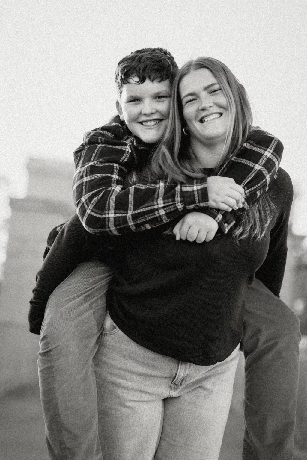 The Garman's family photos taken at BridgeWay Station, Simpsonville: Black and white photo of a smiling woman giving a piggyback ride to a smiling child wearing a plaid shirt outdoors.