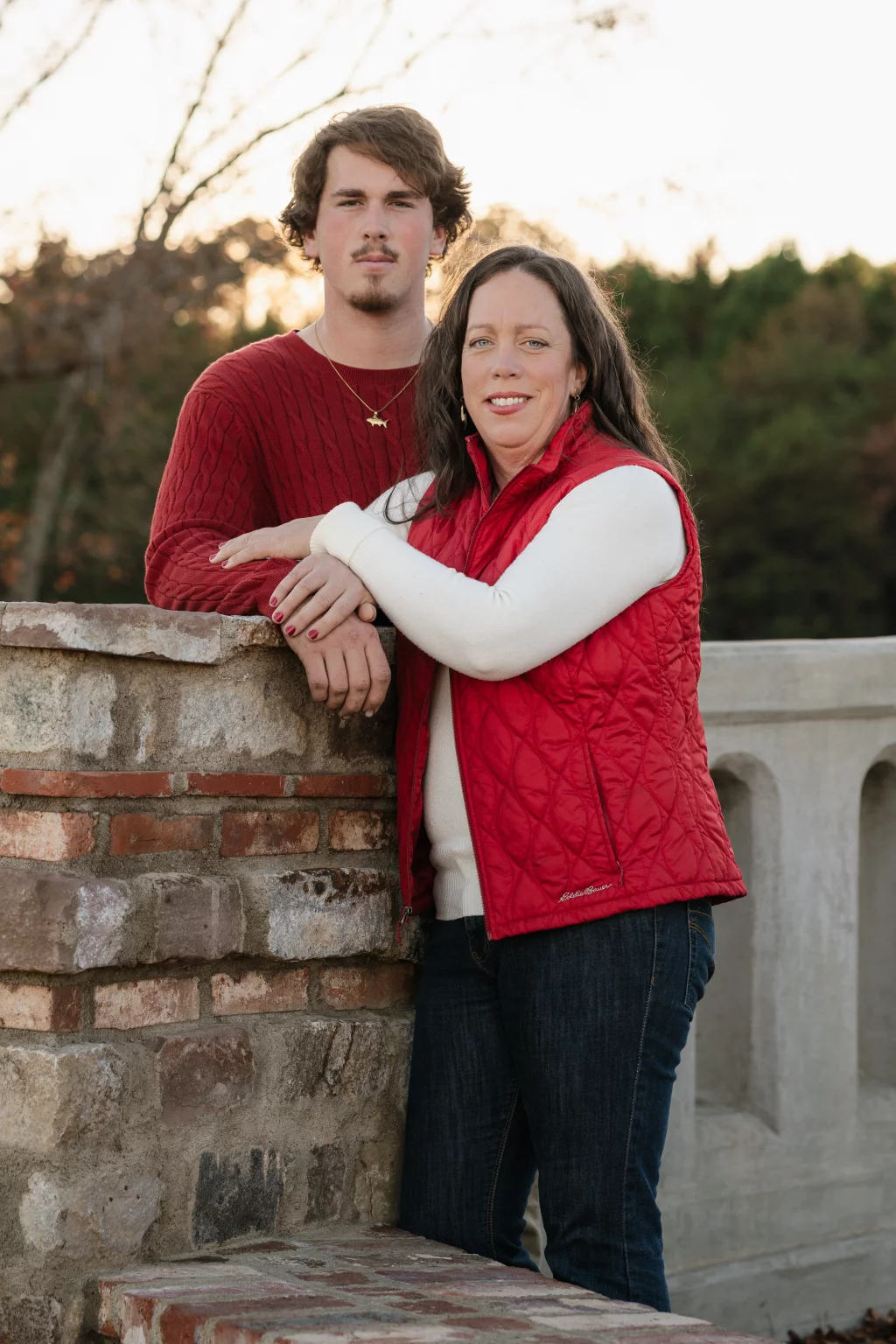 The Garman's family photos taken at BridgeWay Station, Simpsonville: Portrait of a woman in a red quilted vest and white sweater leaning on a stone wall with a young man in a red cable-knit sweater standing behind her outdoors.