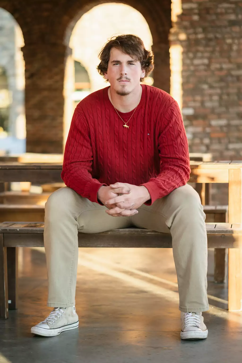 : Young man in a red sweater, beige pants, and white sneakers sitting on a wooden bench with hands clasped, brick arches in the background.