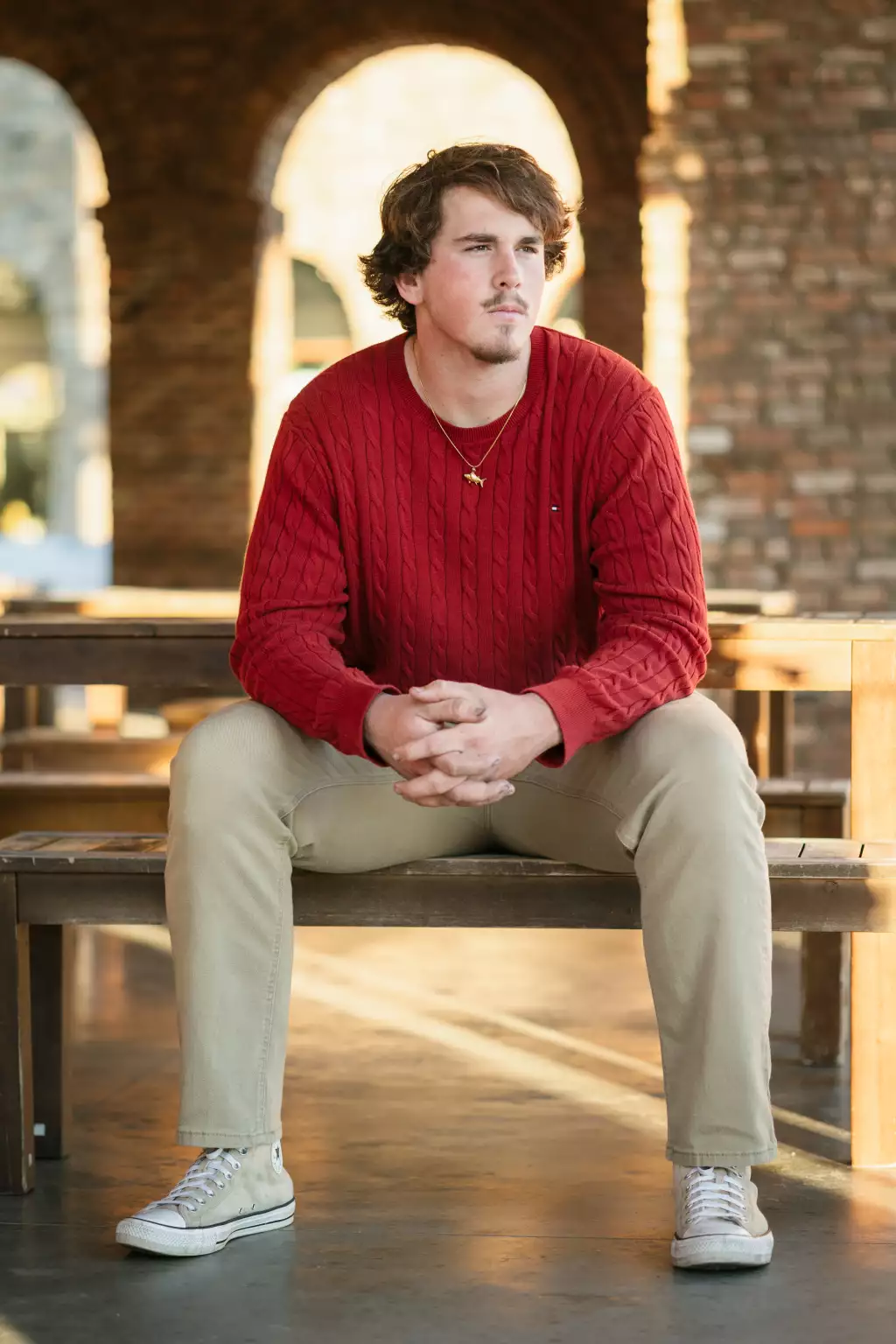 : Young man with curly hair wearing a red sweater and beige pants, sitting on a wooden bench with hands clasped, sunlit arched brick background.