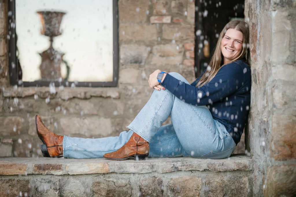 The Garman's family photos taken at BridgeWay Station, Simpsonville: Smiling woman in blue sweater and light jeans sitting on a stone ledge against a stone wall with water droplets in the foreground.