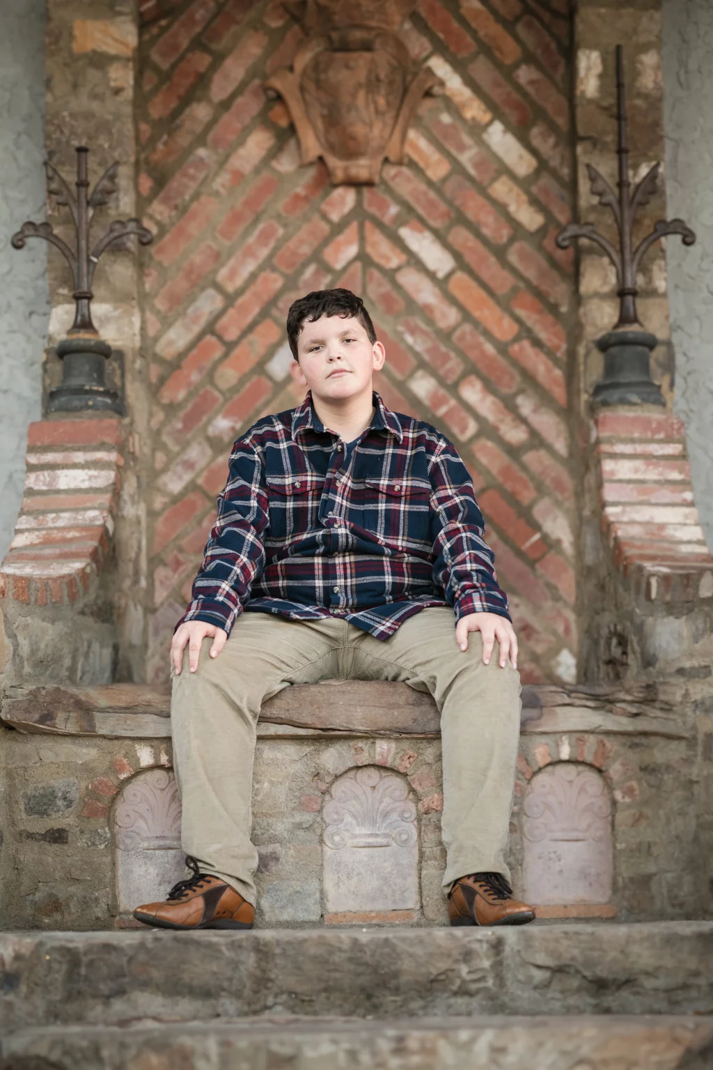 The Garman's family photos taken at BridgeWay Station, Simpsonville: Boy in a plaid shirt and khaki pants sitting on a carved stone bench with a decorative brick and stone wall behind him.