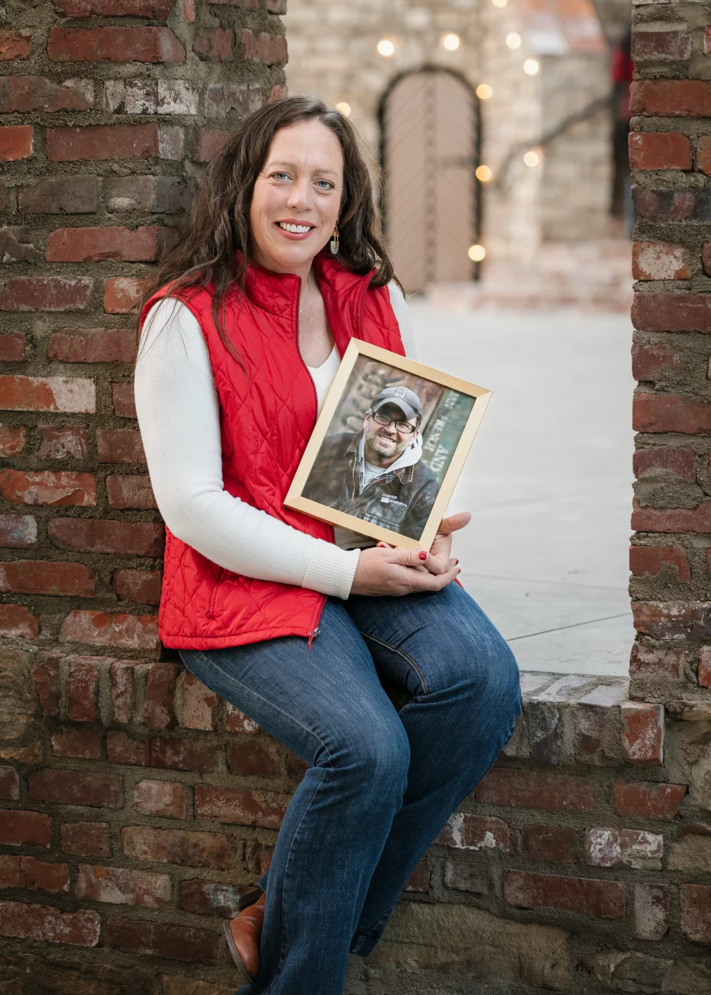 The Garman's family photos taken at BridgeWay Station, Simpsonville: Woman in a red vest and white long-sleeve shirt sitting on a brick ledge, holding a framed photo of a man wearing glasses, a baseball cap, and a jacket.