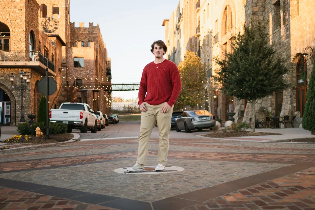 The Garman's family photos taken at BridgeWay Station, Simpsonville: Young man in a red sweater and beige pants standing on a patterned brick street in an urban area with stone buildings and parked cars in the background.