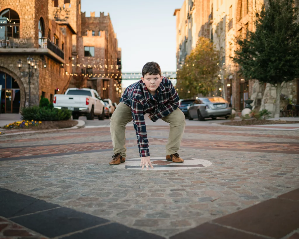 The Garman's family photos taken at BridgeWay Station, Simpsonville: Boy crouching on a cobblestone street in a small urban plaza at sunset, with buildings, cars, and string lights in the background.