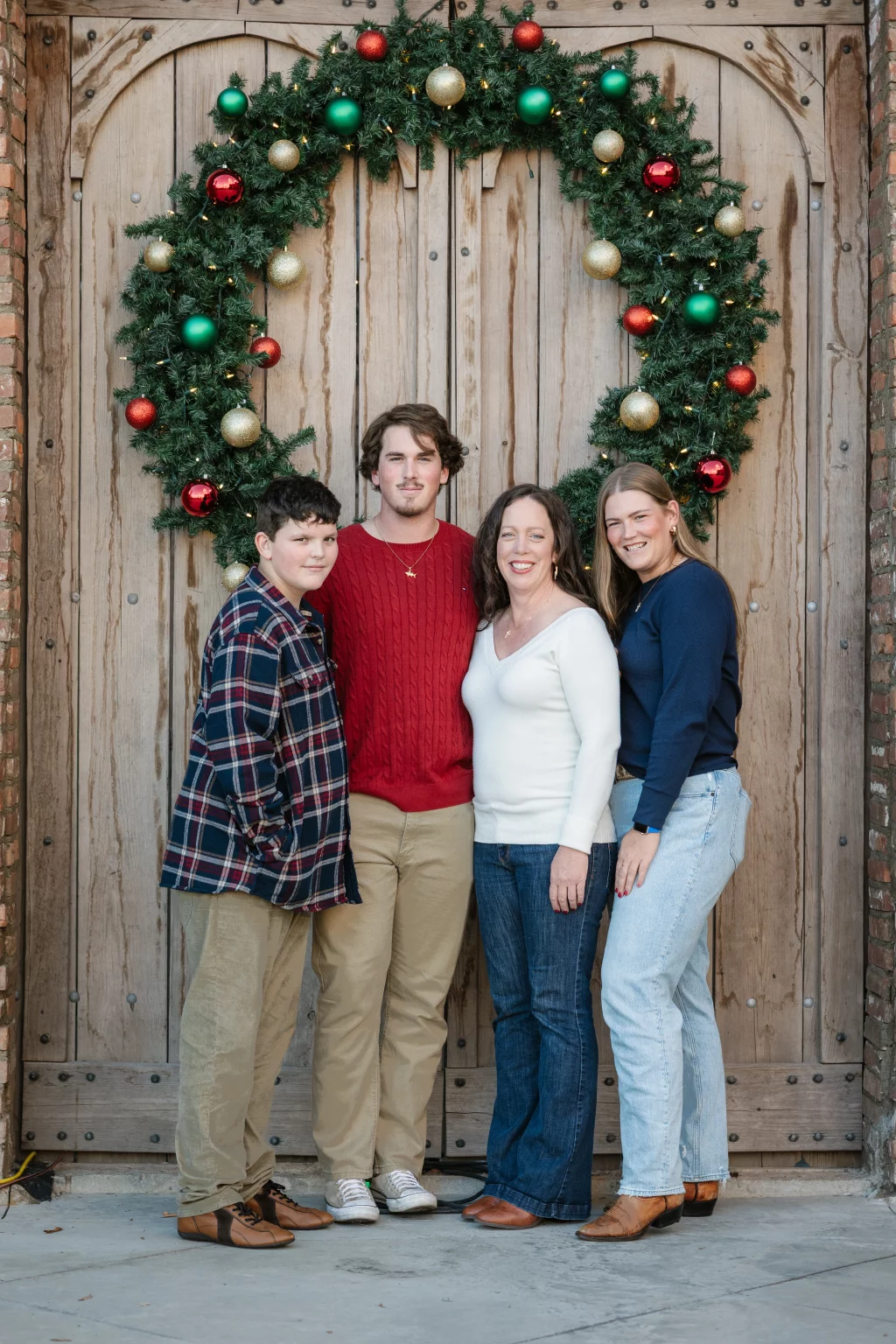 The Garman's family photos taken at BridgeWay Station, Simpsonville: Four people standing in front of a large wooden door adorned with a festive Christmas wreath decorated with red, green, and gold ornaments.