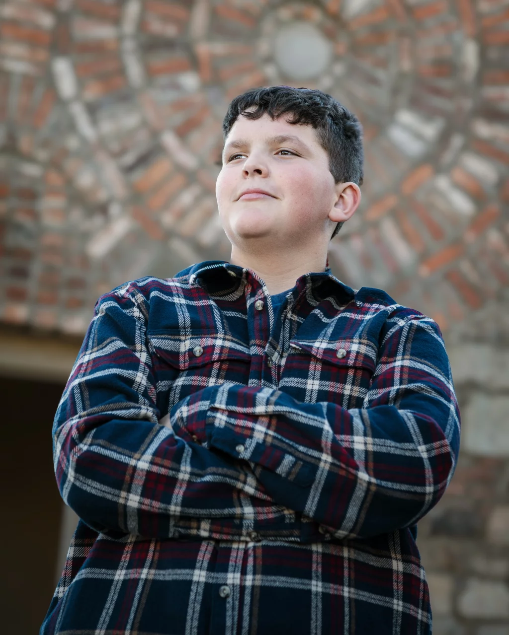 The Garman's family photos taken at BridgeWay Station, Simpsonville: Teenage boy wearing a blue and red plaid shirt standing with crossed arms in front of a patterned brick wall.
