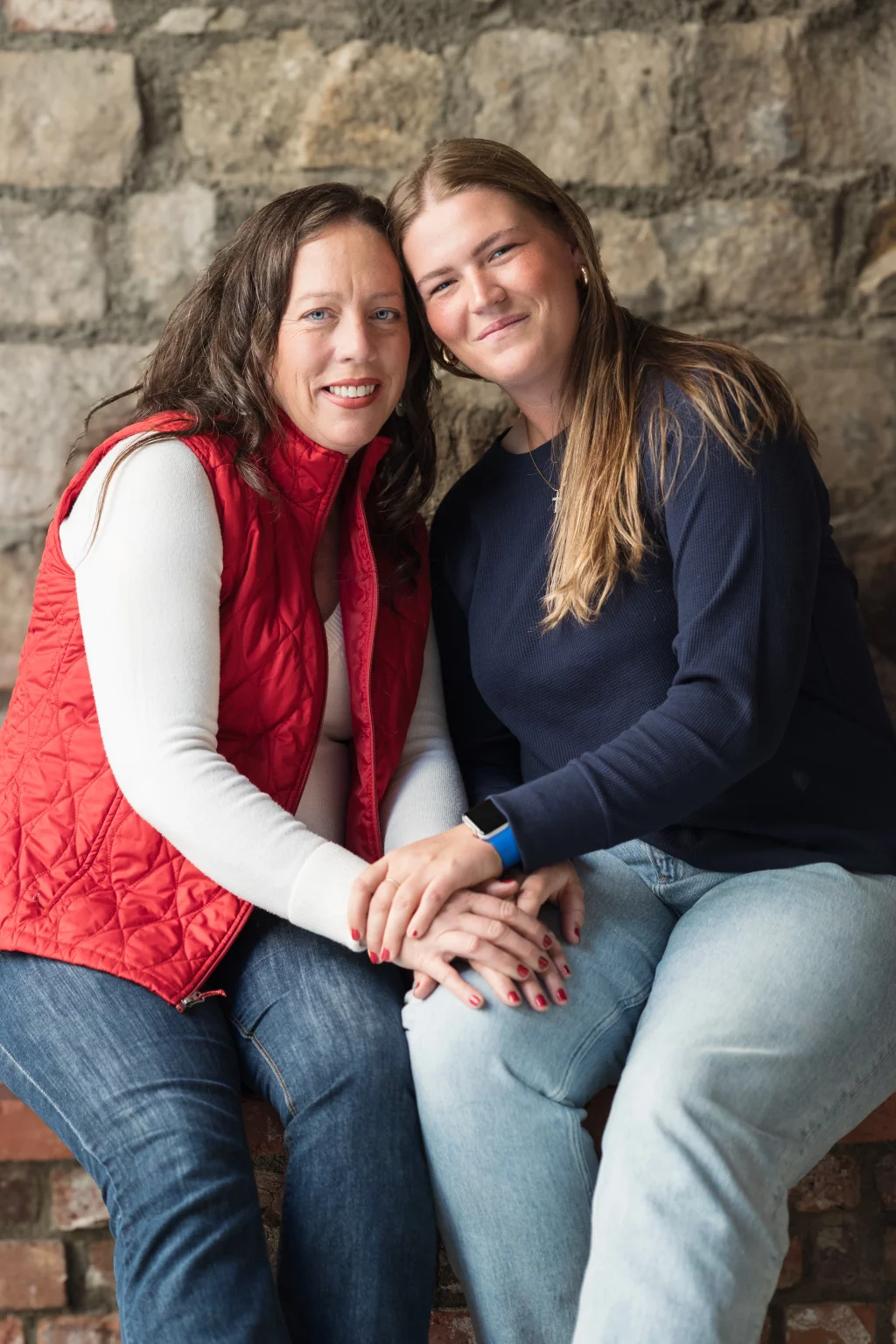 The Garman's family photos taken at BridgeWay Station, Simpsonville: Two women sitting close together against a stone wall, holding hands and smiling at the camera.