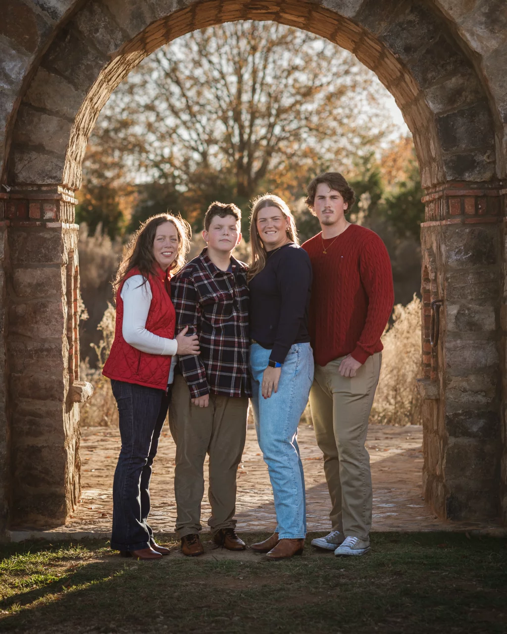 The Garman's family photos taken at BridgeWay Station, Simpsonville: Four people standing together under a stone archway outdoors with trees and sunlight in the background.