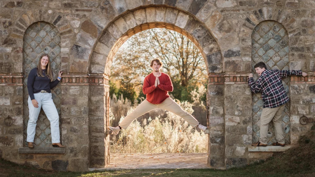 The Garman's family photos taken at BridgeWay Station, Simpsonville: Three people posing on a stone archway structure outdoors, one person in a red sweater jumping with legs spread in the center arch, two others standing in smaller side alcoves, one smiling and one playfully leaning.