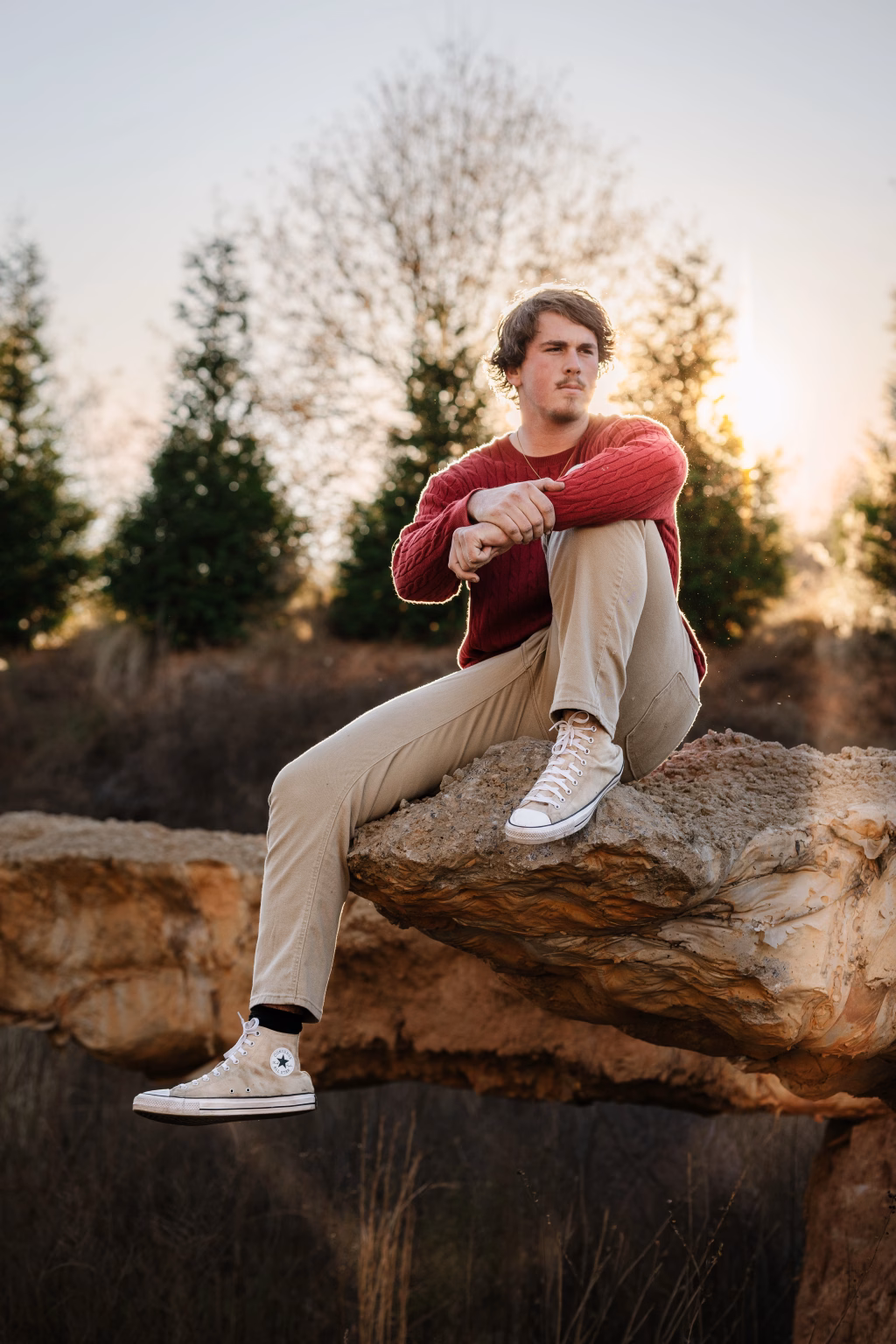 The Garman family photos taken at BridgeWay Station, Simpsonville: Young man in a red sweater and beige pants sitting thoughtfully on a large rock ledge outdoors with trees and sunlight in the background.