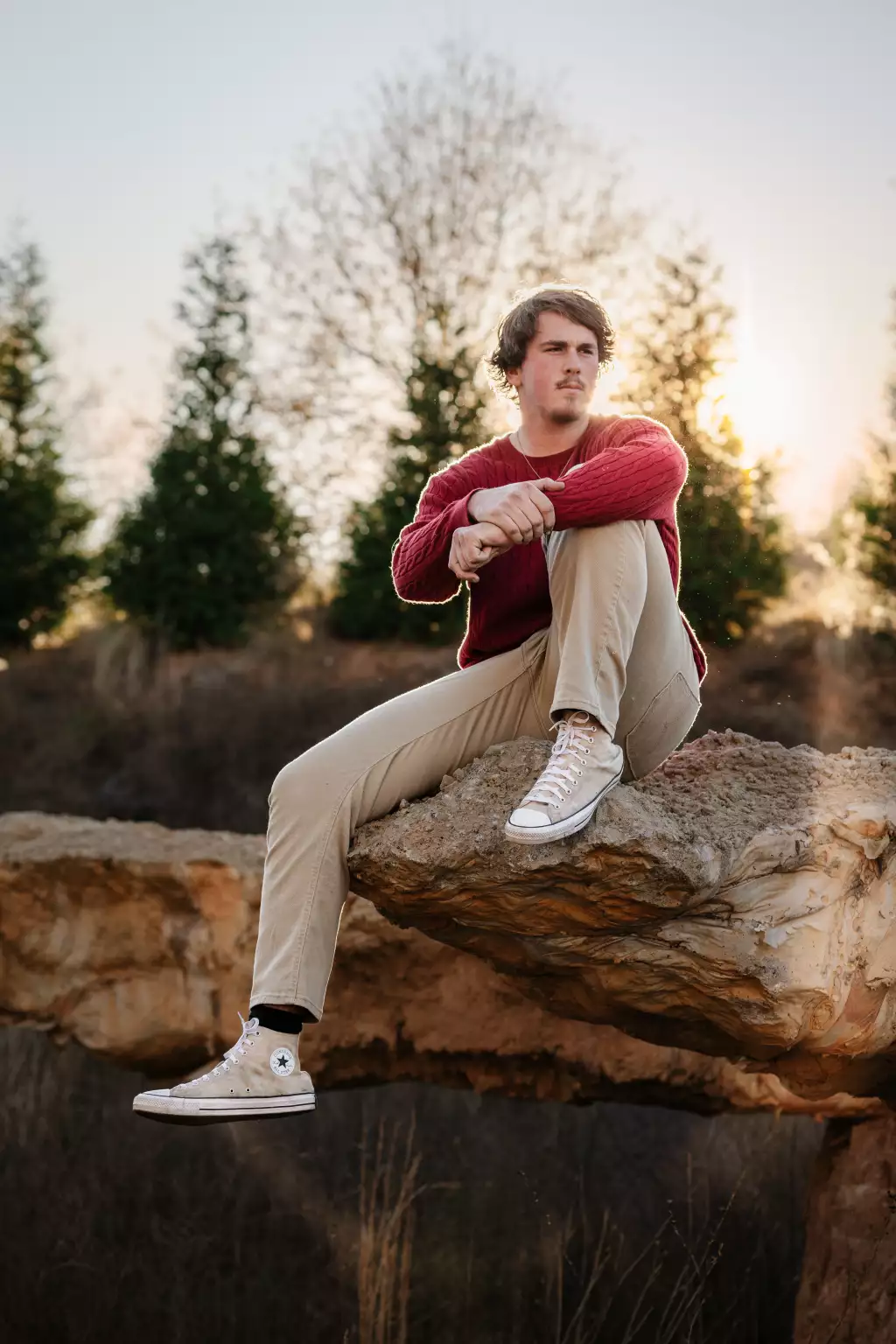 : Young man in a red sweater and beige pants sitting thoughtfully on a large rock ledge outdoors with trees and sunlight in the background.