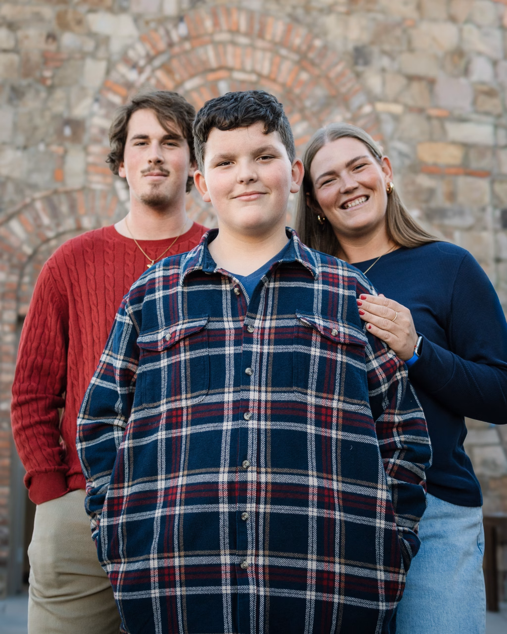 The Garman family photos taken at BridgeWay Station, Simpsonville: Portrait of three people standing closely in front of a stone wall with a brick arch; a smiling boy in a blue and red plaid shirt is in front, with a young man in a red sweater and a smiling woman in a blue top behind him.