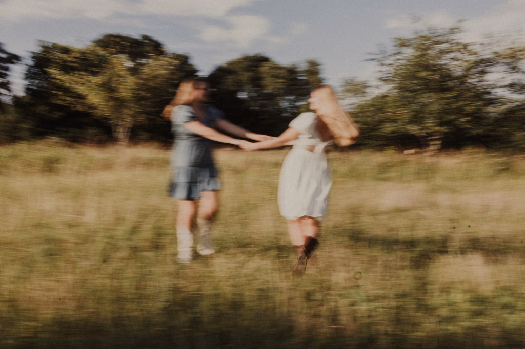 Grace and Morgan's photos taken at Conestee Park, Greenville, SC: Two people holding hands and spinning around in a grassy field with trees in the background, image is blurred showing motion.