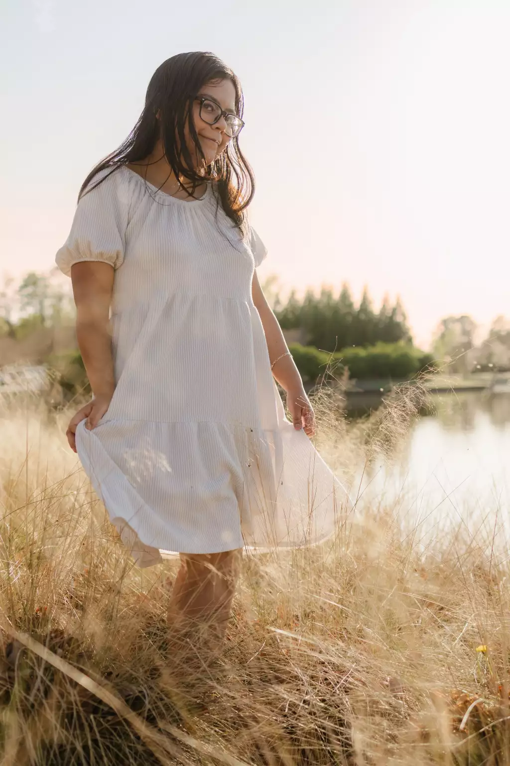 : Young woman with long dark hair and glasses wearing a light-colored dress, standing in tall grass near a body of water with sunlight softly illuminating the scene.