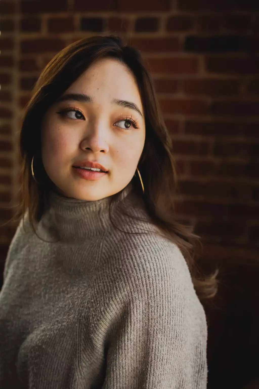 : Portrait of a young woman with long dark hair wearing a beige knitted turtleneck sweater and gold hoop earrings, looking slightly to her left against a dark brick wall background.