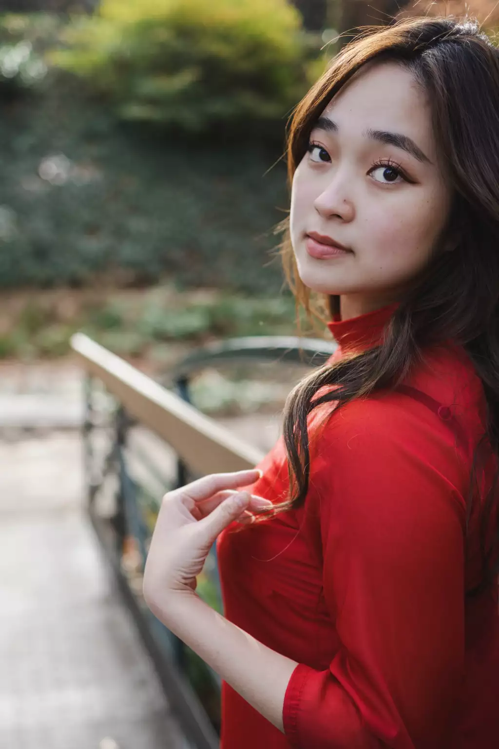 : Portrait of a young woman with long dark hair wearing a red top, standing outdoors near a railing, looking over her shoulder toward the camera with a soft expression.