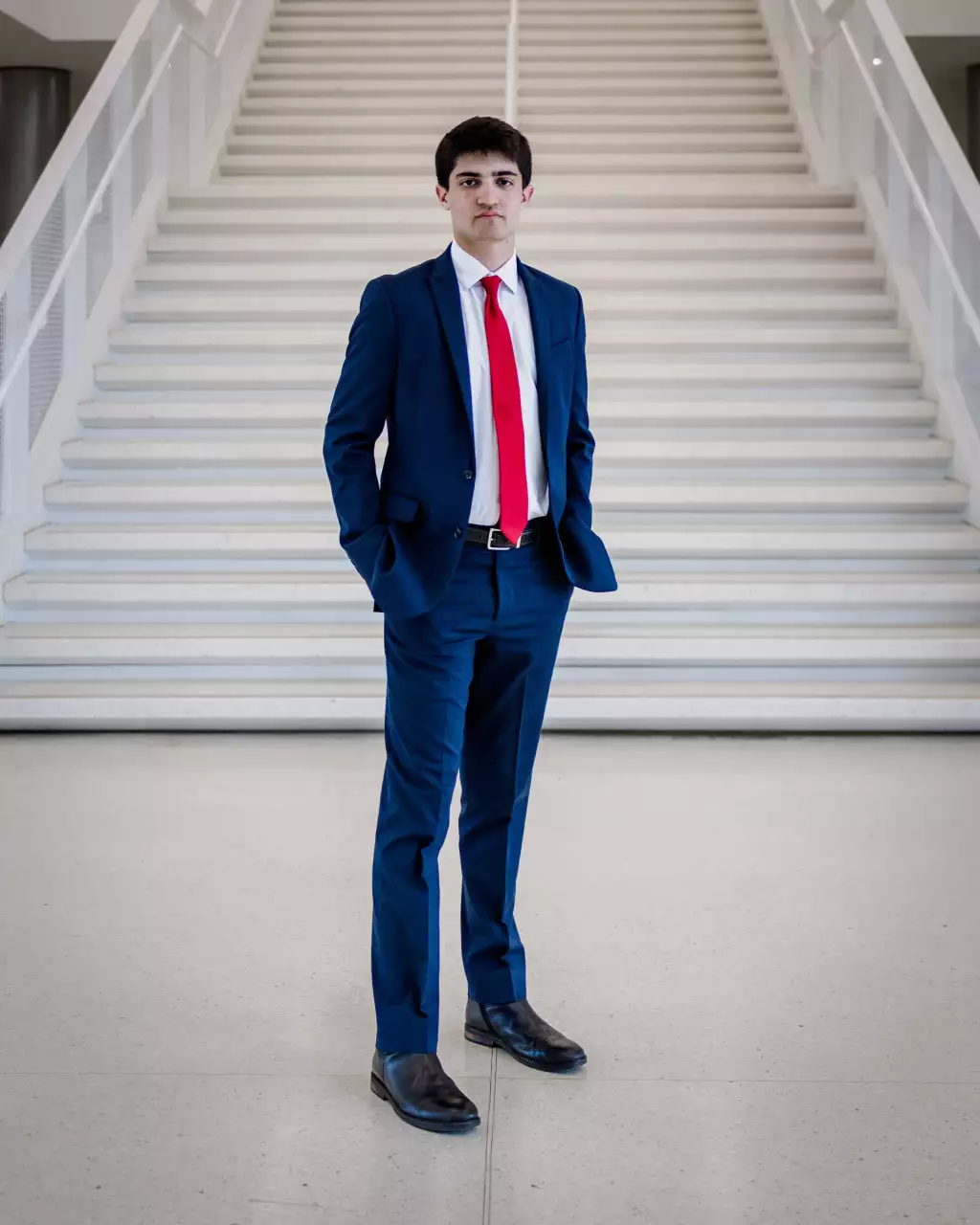 : Young man standing in front of a wide white staircase, wearing a dark blue suit, white shirt, red tie, and black shoes, with hands in his pockets.