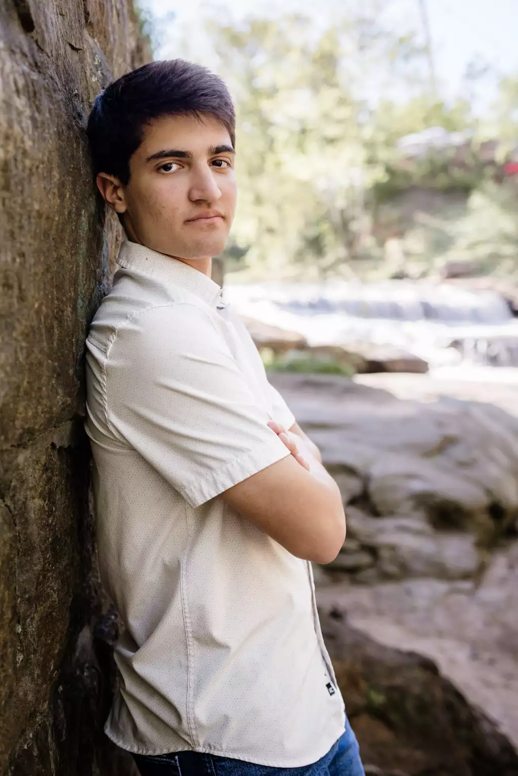 : Young man with short dark hair leaning against a textured rock wall outdoors, wearing a light short-sleeve button-up shirt and jeans, with a blurred background of rocks, greenery, and flowing water.
