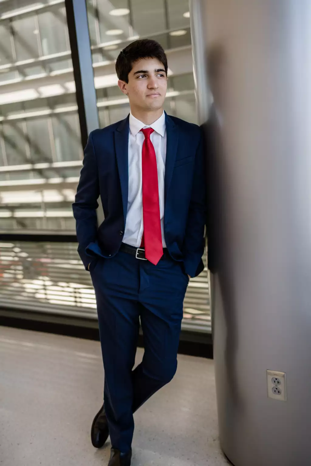 : Young man in a navy blue suit with a red tie, white shirt, and black shoes, standing indoors leaning against a large metallic column with hands in pockets, looking slightly to the side.