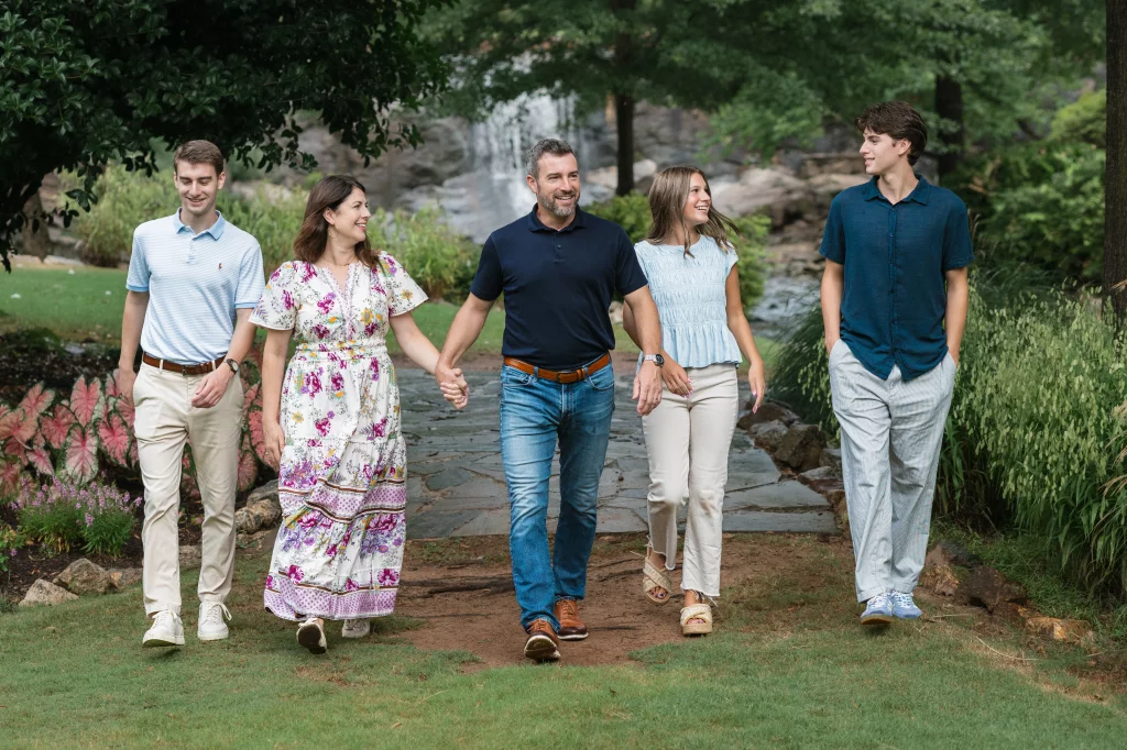: Family of five walking hand in hand on a garden path, surrounded by greenery and flowers, with a waterfall in the background.