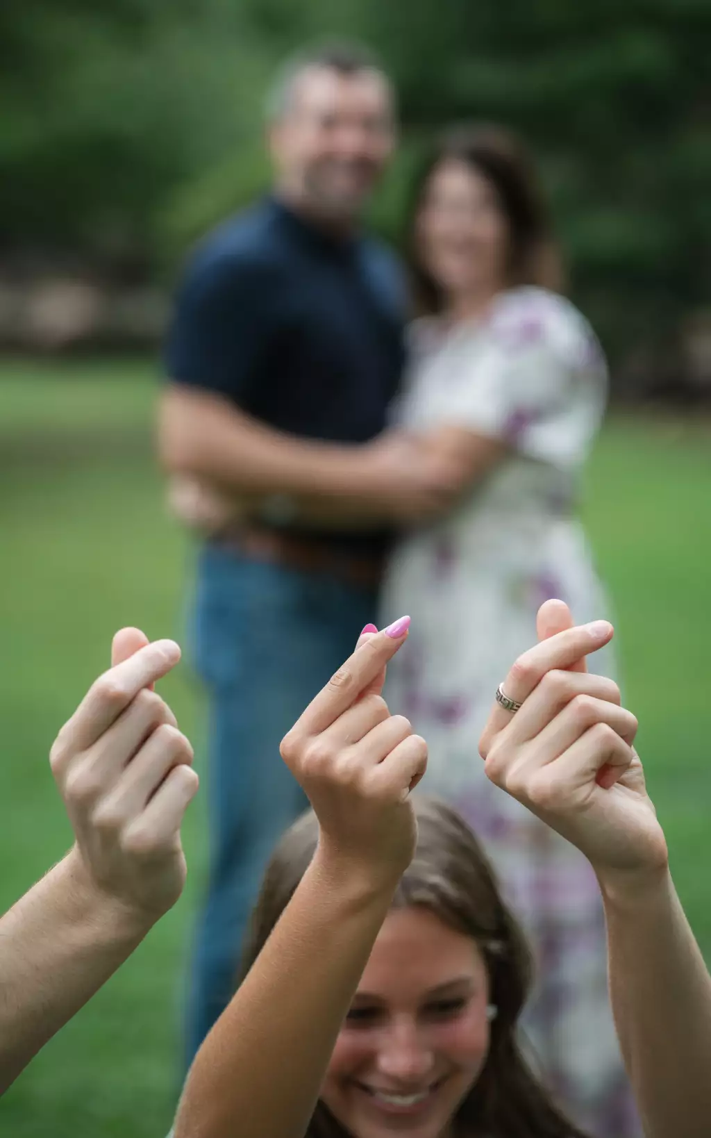 : Close-up of three hands making Korean finger heart gestures in the foreground, with a smiling young woman behind them and a blurred couple embracing in the background outdoors on green grass.