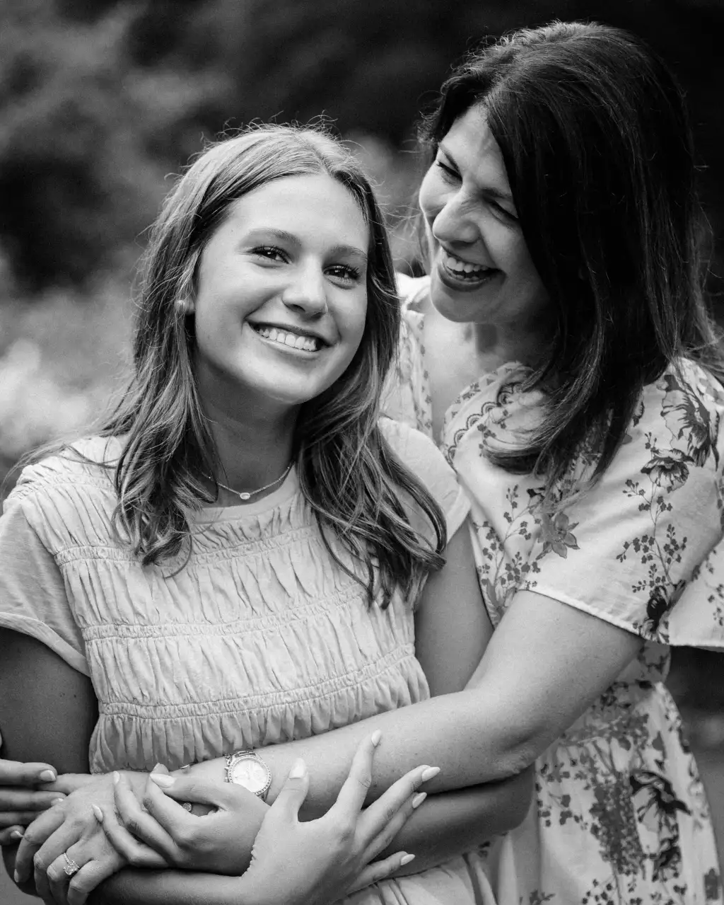 : Black-and-white photo of a smiling young woman embraced from behind by another woman who is laughing, both outdoors with blurred greenery in the background.
