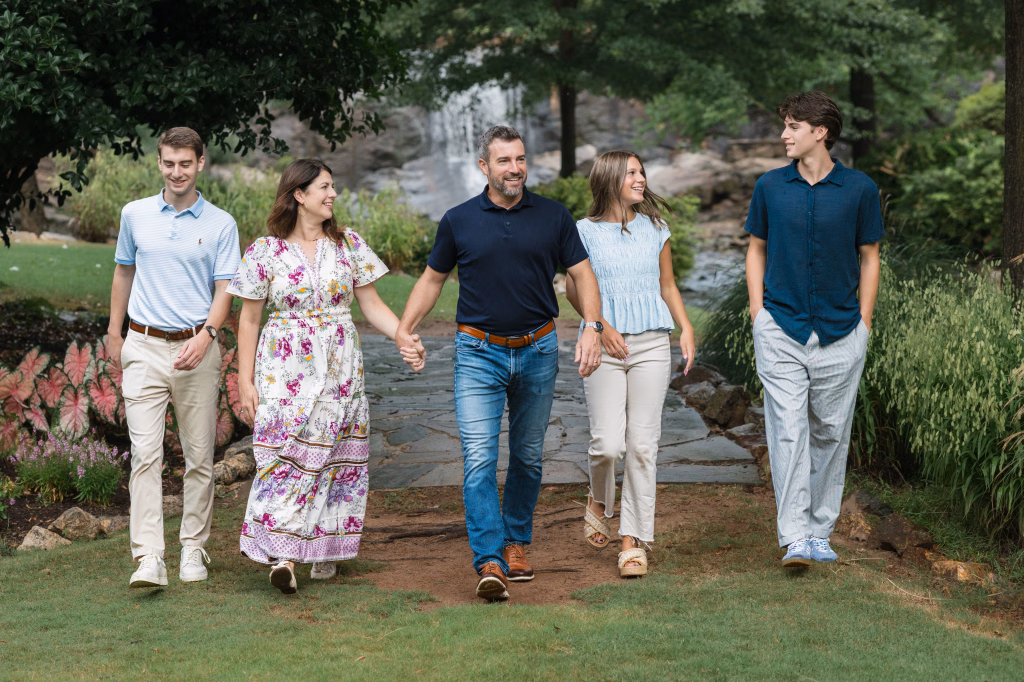 The Hotchkin family photos taken at Rock Quarry Garden, Greenville, SC: Five people&mdash;two adults and three teenagers&mdash;walking together on a garden path, surrounded by greenery and flowers, with a waterfall in the background. The adults in the center are holding hands and all are smiling and looking at each other.
