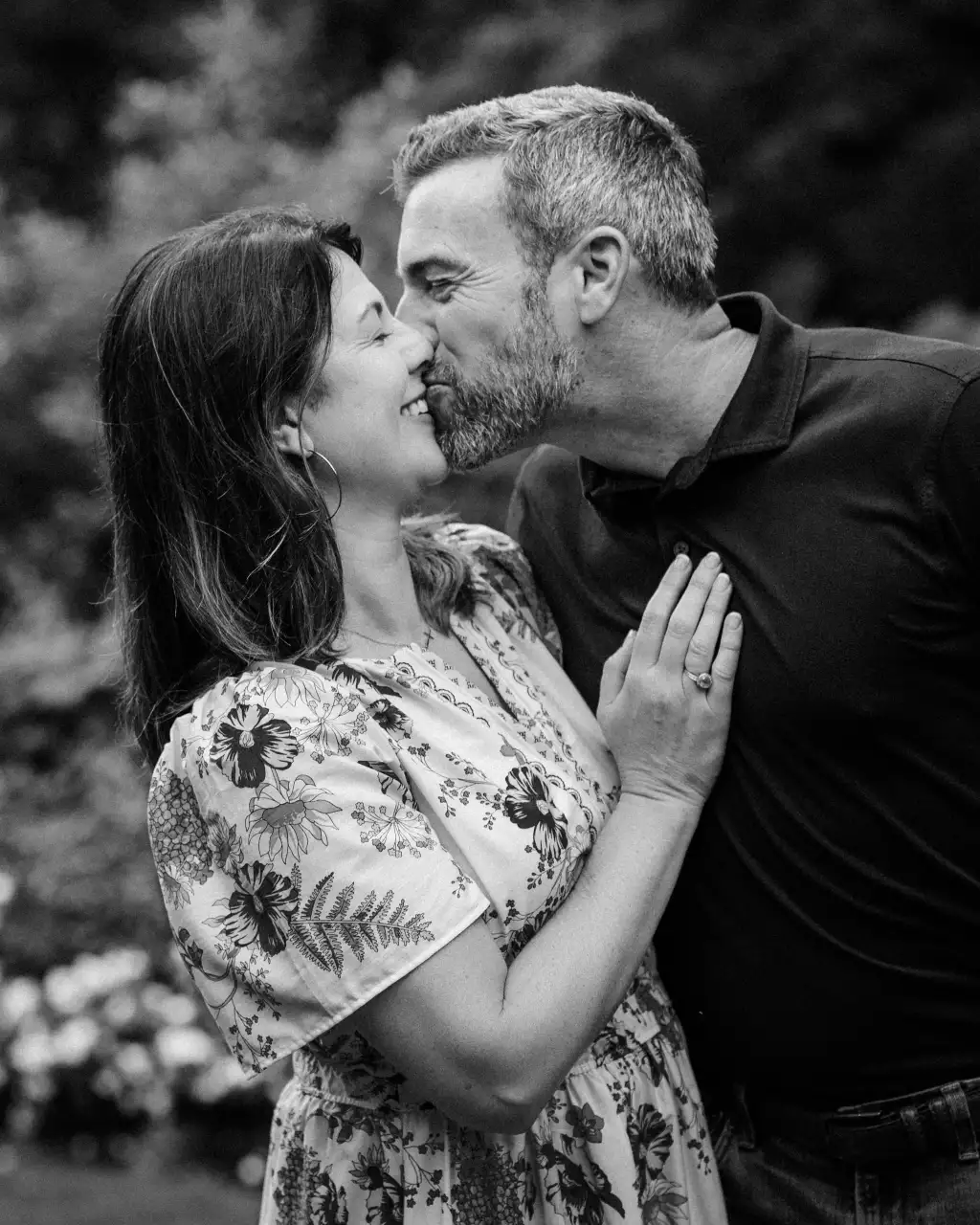 : Black and white photo of a couple sharing a tender kiss, the woman smiling with eyes closed, wearing a floral dress and hoop earrings, and the man leaning in wearing a dark polo shirt with short hair and beard. The woman&rsquo;s hand is gently resting on the man&rsquo;s chest, showing a ring. The background is blurred greenery.