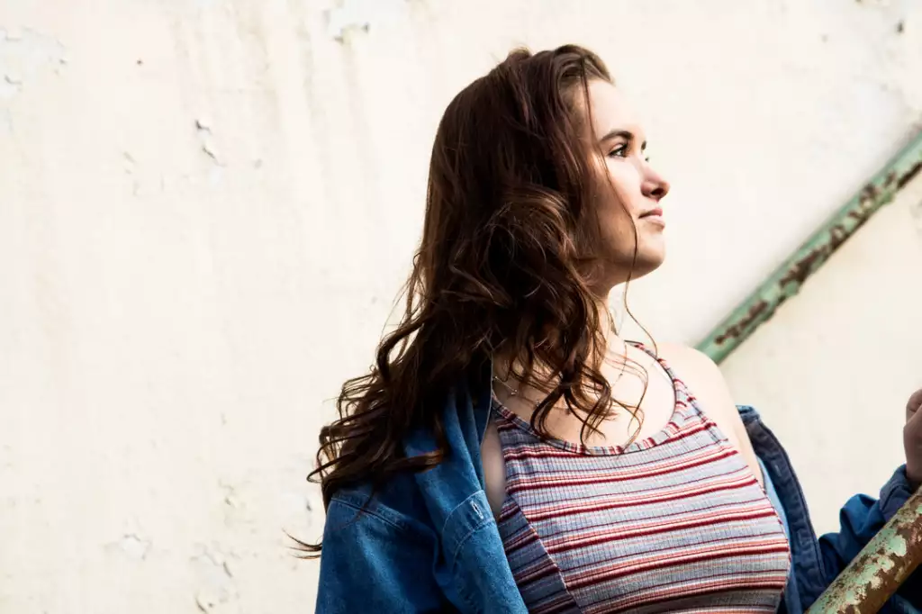 Katelynd's senior photos at Taylor's Mill: Young woman with long wavy brown hair wearing a striped tank top and a denim jacket, looking to the right, standing next to a rusty green metal railing against a textured beige wall.