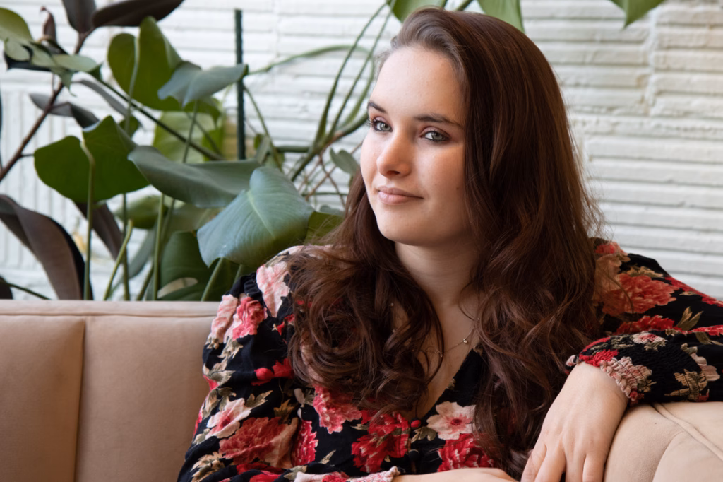 Katelynd's senior photos at Taylor's Mill: Woman with long brown hair wearing a black floral blouse, sitting on a beige couch with green leafy plants and a white brick wall in the background.