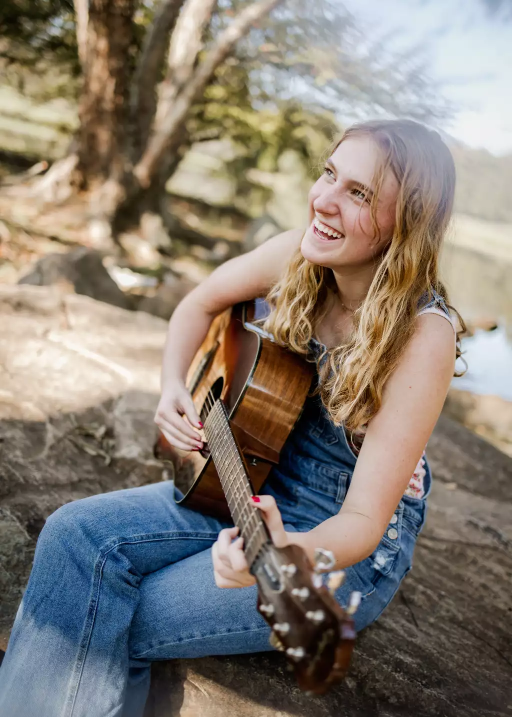 : Young woman with long wavy blonde hair sitting outdoors on a rock, smiling and playing an acoustic guitar, wearing blue denim overalls.