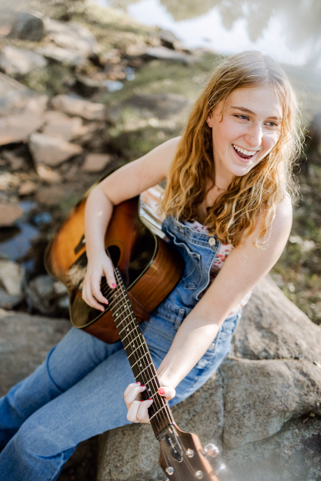 Lena's senior photo taken at the Milliken Arboretum: Young woman with long blonde hair sitting on a rock near water, smiling and playing an acoustic guitar, wearing denim overalls and a floral top.