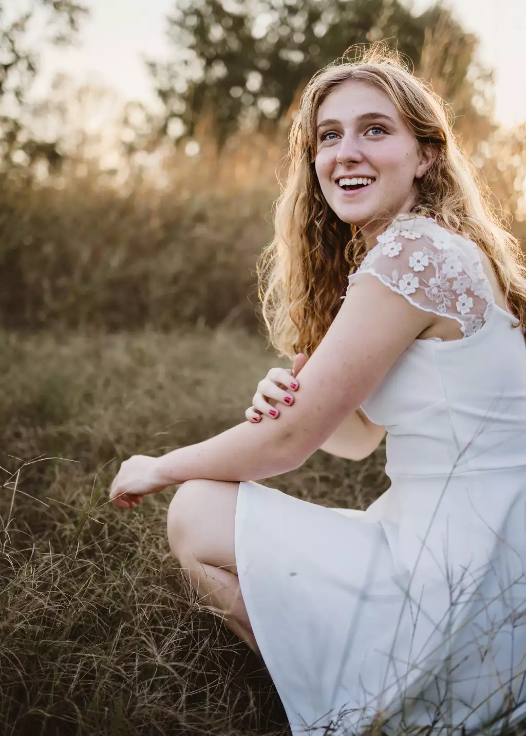 : Young woman with wavy blonde hair wearing a white dress with floral lace sleeves, smiling and crouching in tall grass during golden hour.