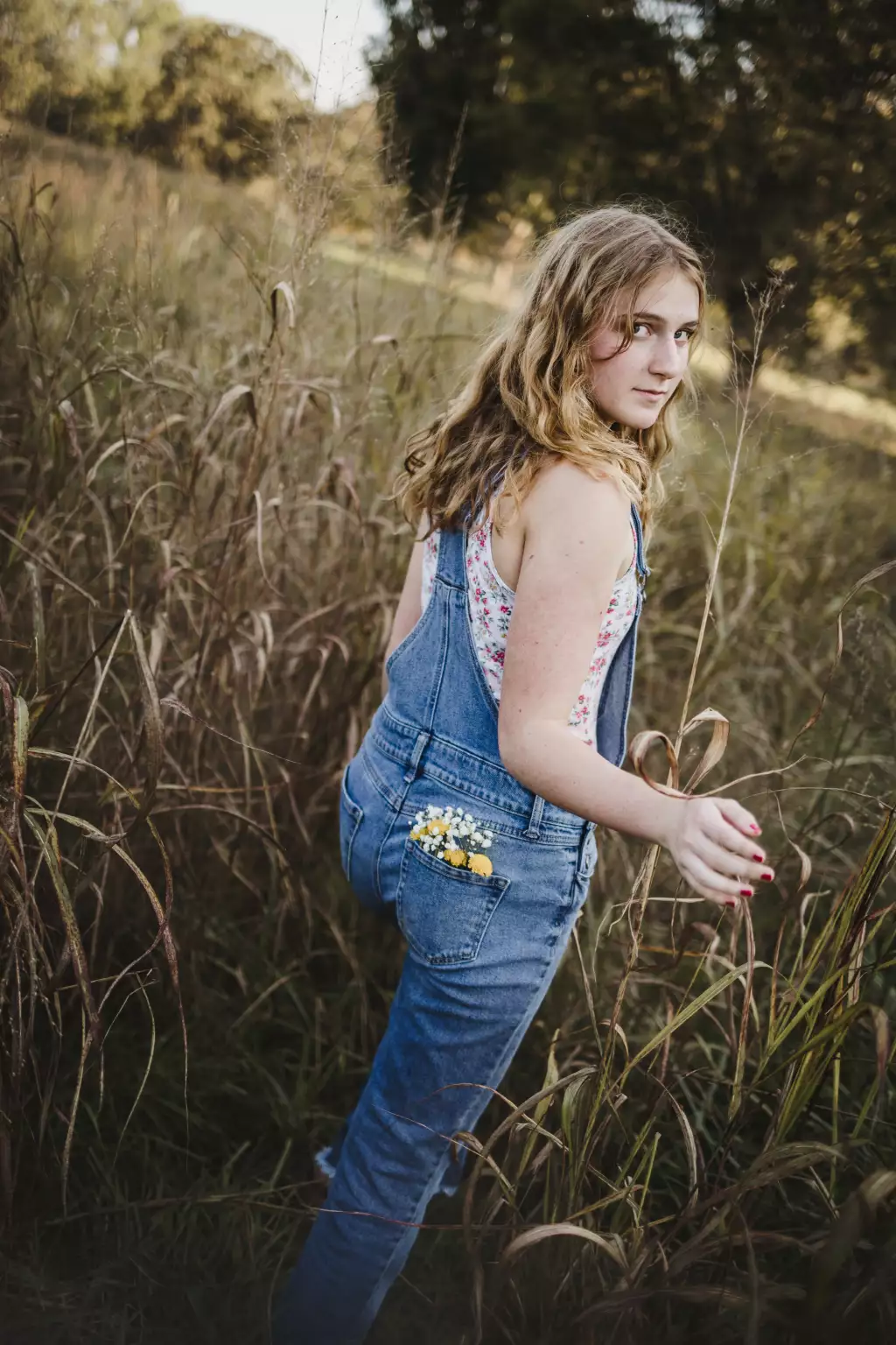 : Teenage girl with wavy blonde hair wearing denim overalls and a floral top standing in a tall grassy field, looking back over her shoulder, with small yellow and white flowers tucked in her back pocket.