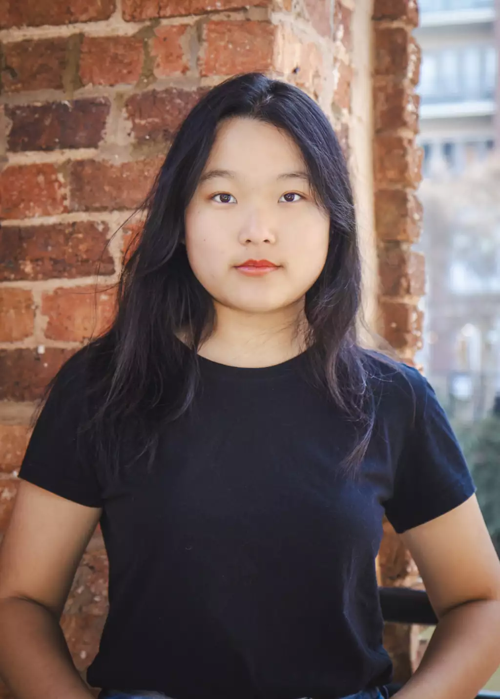 Lily's senior photos taken downtown Greenville, SC: Portrait of a young woman with long black hair wearing a black t-shirt, standing in front of a red brick wall, looking directly at the camera with a neutral expression.