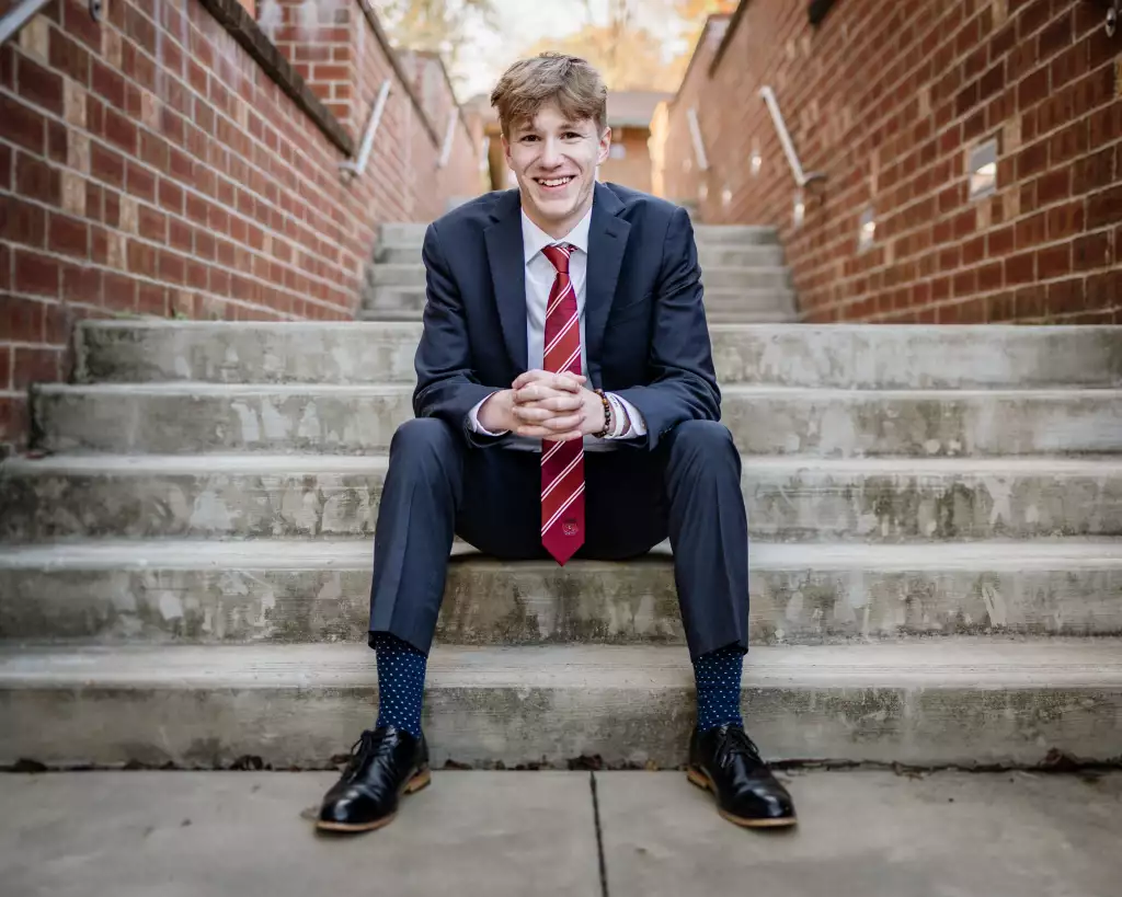 : Young man in a dark suit with a red striped tie sitting on concrete steps between brick walls, smiling with hands clasped.
