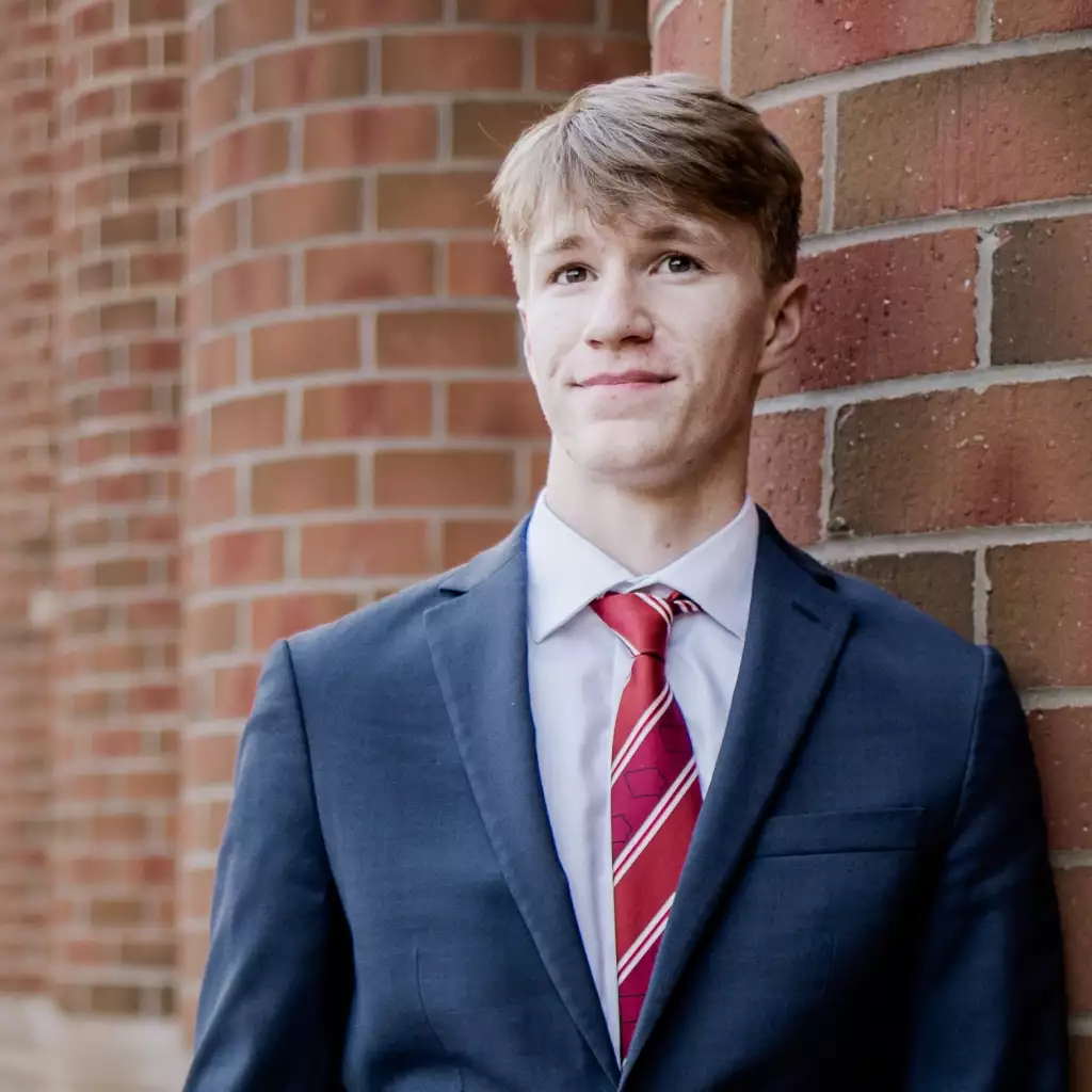 Mac's senior photos: Young man in a navy suit, white shirt, and red striped tie, leaning against a brick wall, looking slightly upward with a neutral expression.