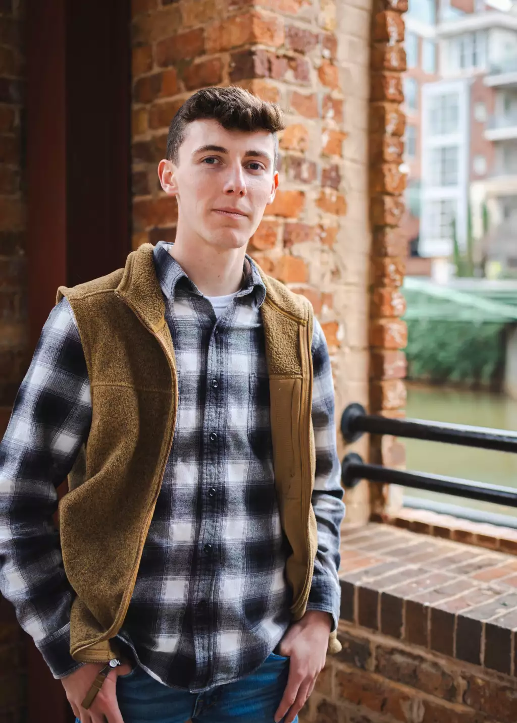 : Young man wearing a brown fleece vest over a black and white plaid shirt, standing with hands in pockets in front of a brick wall and railing near a body of water.