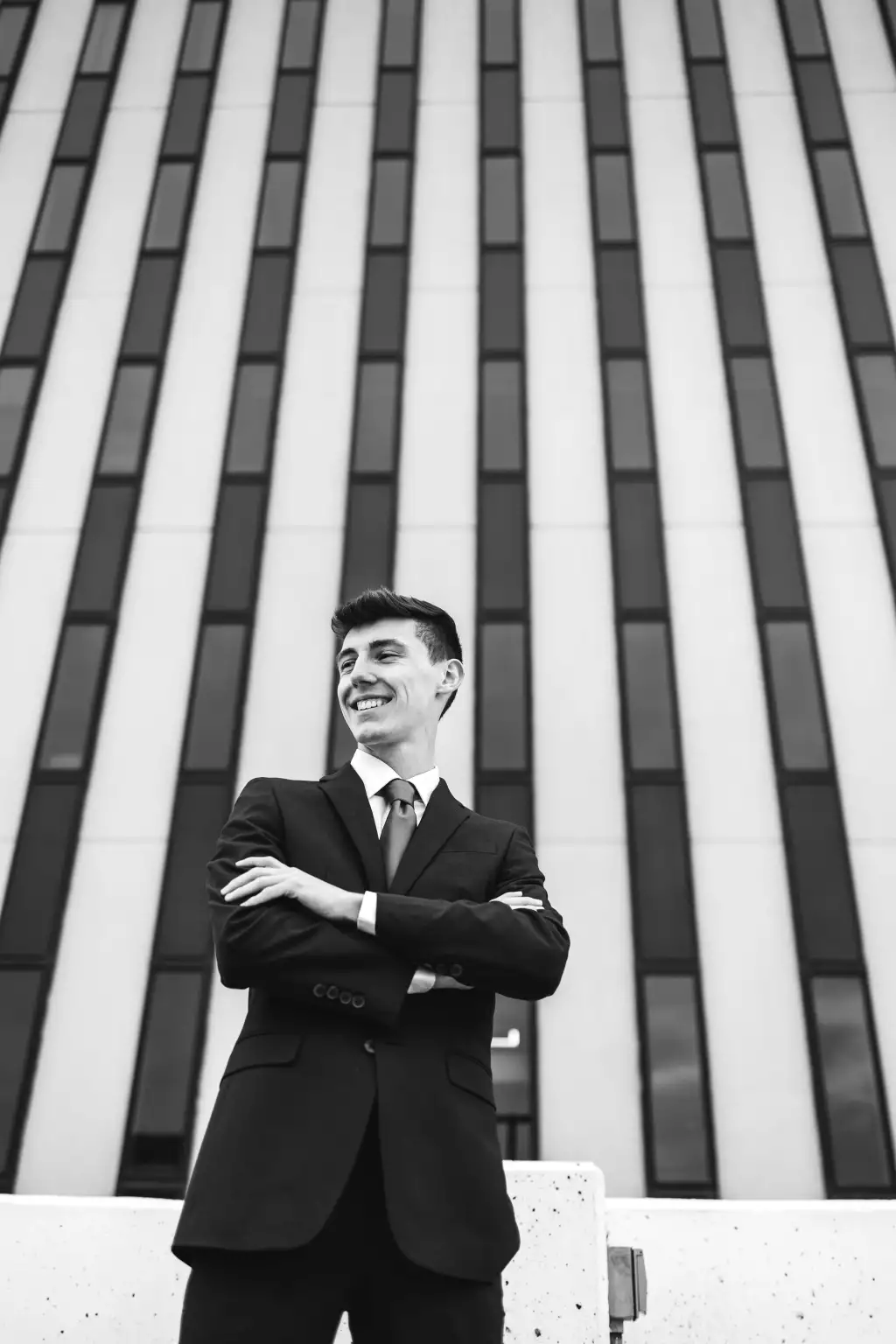 : Black and white photo of a smiling young man in a suit and tie, standing with arms crossed in front of a tall building with vertical rectangular windows.