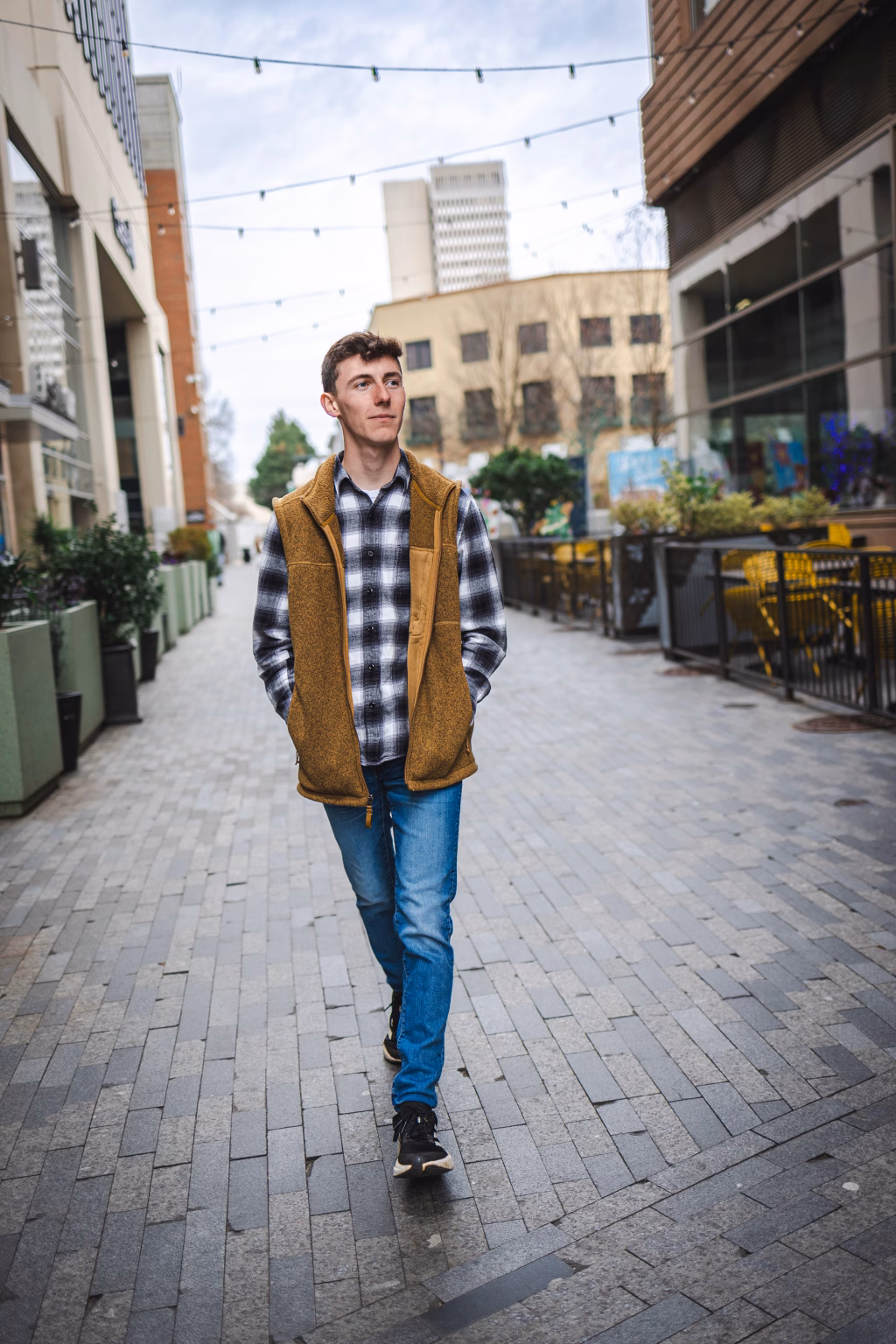 Madigan's senior photos taken in downtown Greenville, SC: Young man walking down a city alleyway with hands in pockets, wearing a mustard vest over a black-and-white checkered shirt, blue jeans, and black sneakers.