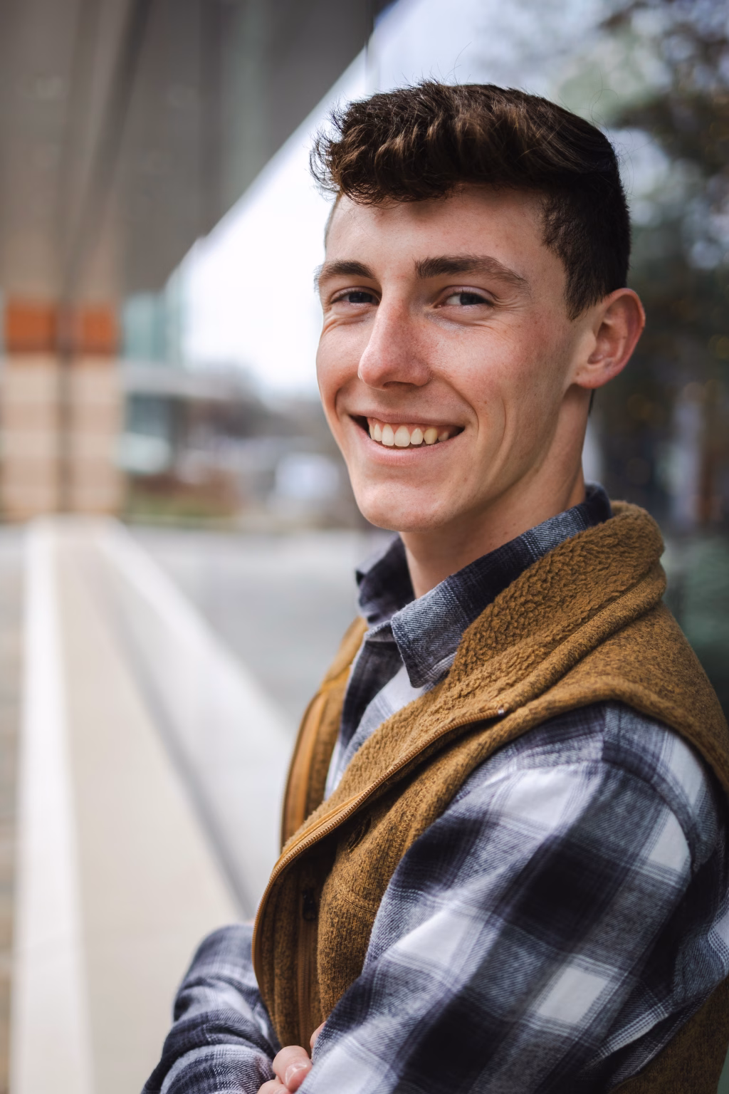 Madigan's senior photos taken in downtown Greenville, SC: Smiling young man with short brown hair wearing a brown fleece vest over a black and white plaid shirt, standing outdoors with arms crossed in front of a building with glass windows.