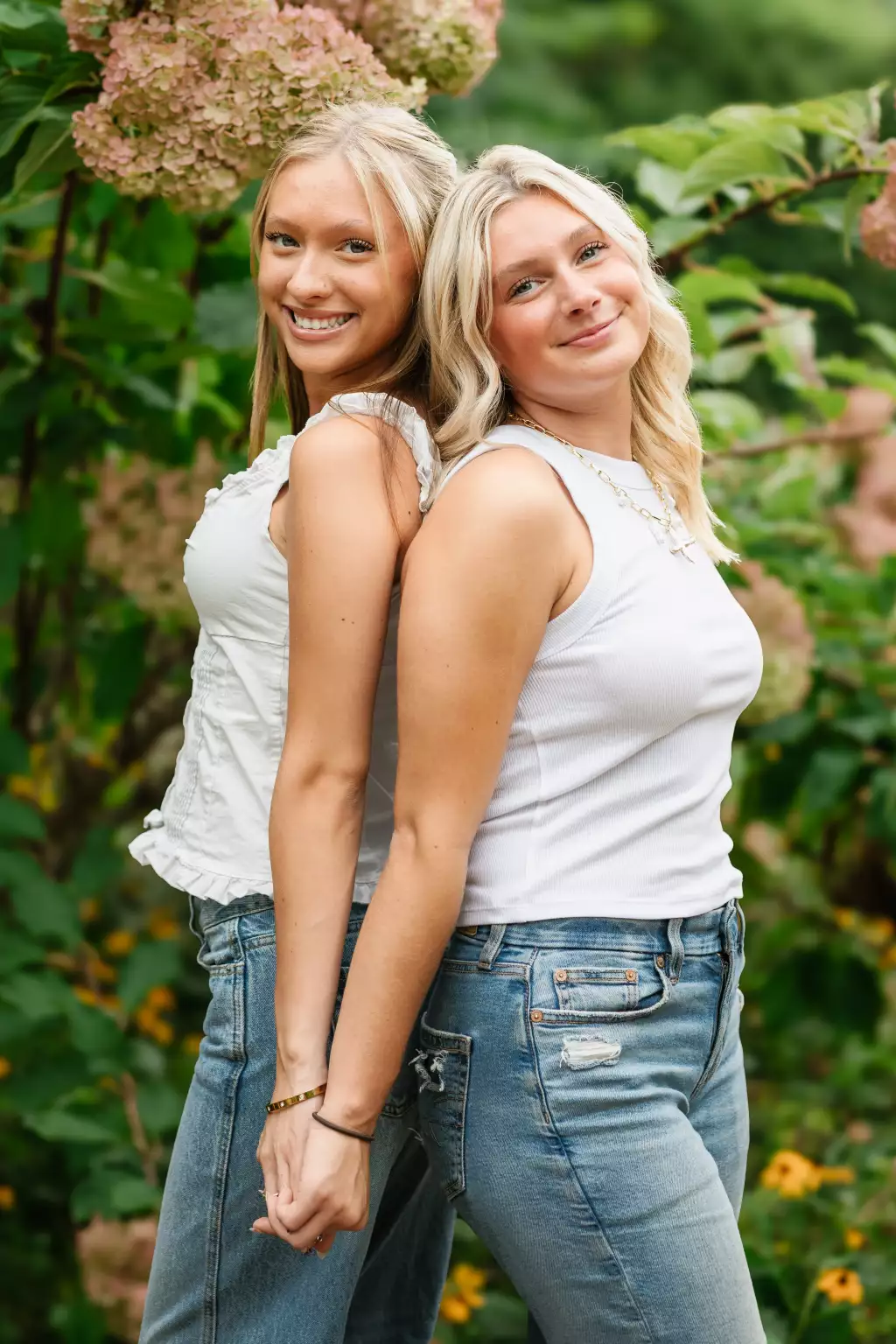 : Two smiling young women standing back-to-back holding hands outdoors with greenery and pink hydrangea flowers in the background.